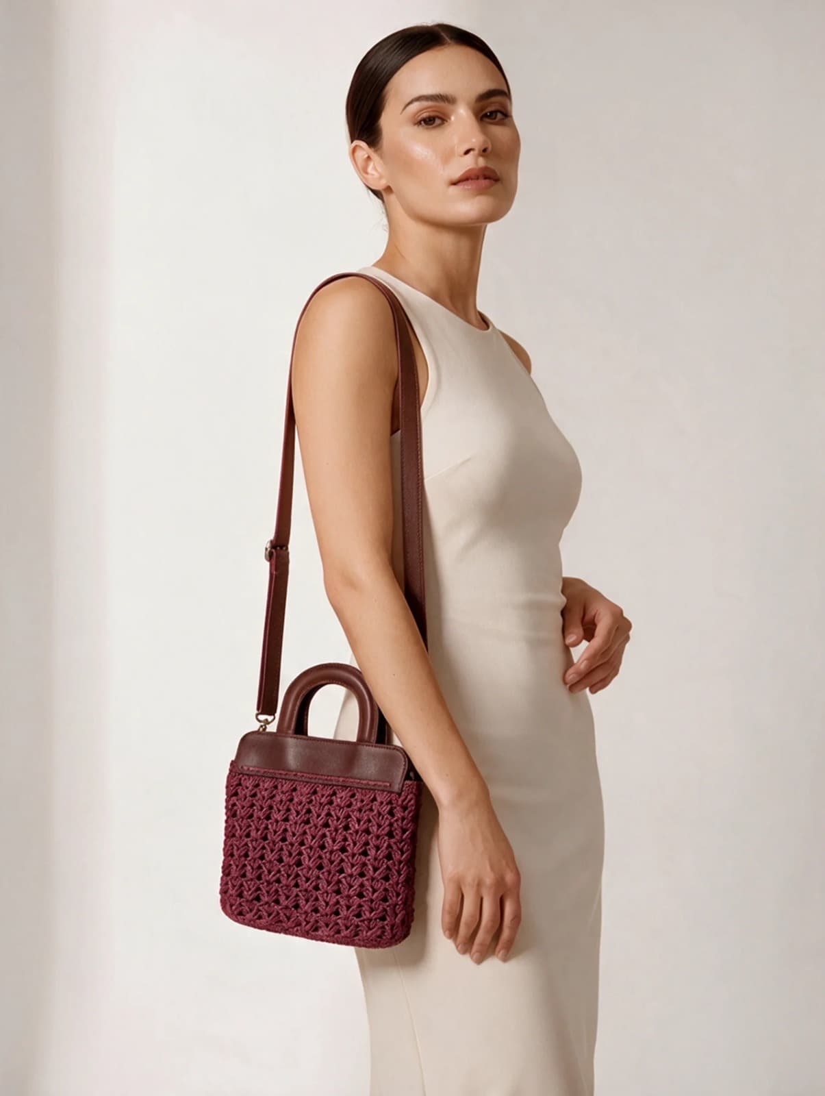 Model in a cream dress carrying the Rete Shoulder Bag, a small burgundy crochet handbag on a long leather strap
