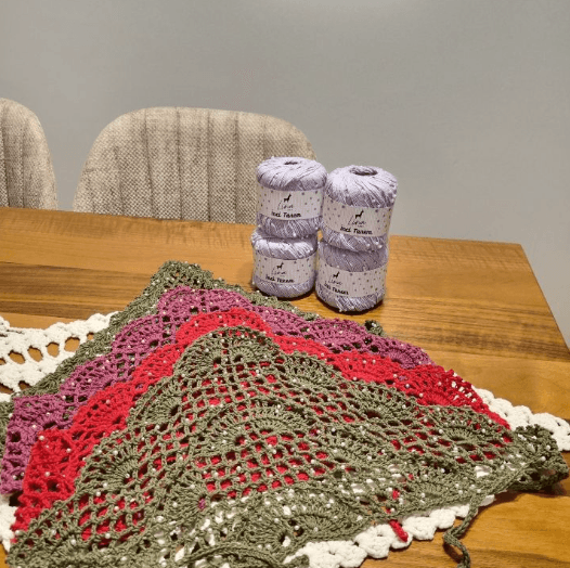 A green, red and pink crochet shawl laid on a table beside lilac yarn cones
