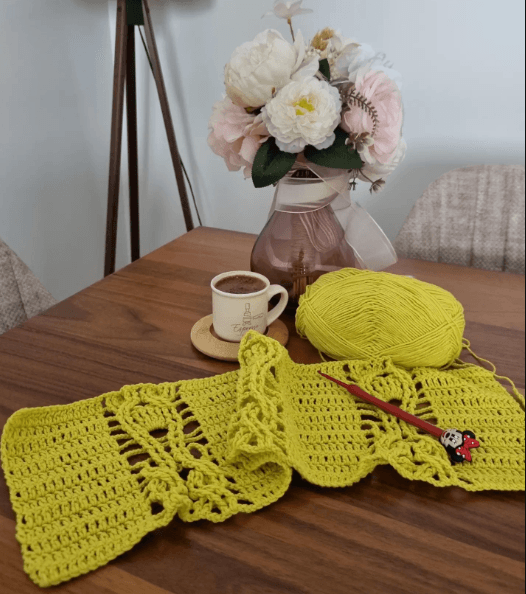 A yellow crochet piece in progress on a table, beside a ball of yarn and flowers